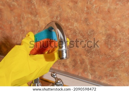 Woman in yellow rubber gloves washes a metal faucet with a sponge at the sink in the kitchen.