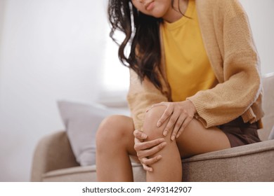 A woman in a yellow shirt is sitting on a couch with her knee propped up. She is in pain and is holding her knee