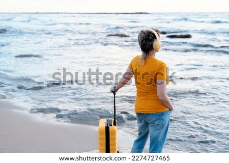 Woman in a yellow shirt with a yellow suitcase and headphone on the seaside on hot sunny day arrived in a tourist town.  Travel  Lifestyle concept