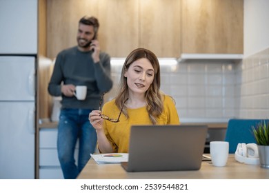 A woman in a yellow sweater focuses on her laptop while a man talks on the phone in the kitchen. The environment is casual and modern, suggesting teamwork and productivity at home. - Powered by Shutterstock