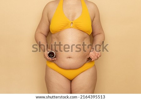 Woman in yellow swimsuit with thick sagging stomach hold jar of body cream in hand, beige background. Slimming, fighting overweight, obesity and cellulite. Plus size people and body positive concept.