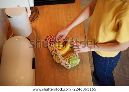 A woman in a yellow T-shirt unpacks an ecological mesh bag with organic products in her kitchen. The concept of reusable packaging