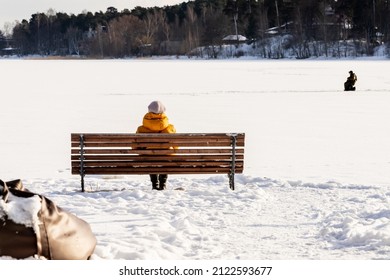 Woman In Yellow Winter Coat Sitting On Her Back On A Wooden Bench Watching Frozen Lake And Male Ice Fisherman In Distance At Winter. Rear View To Resting Woman At Sunny But Cold Winter Day.