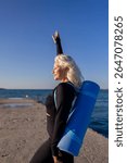 Woman Yoga Mat Beach - Blonde woman in black leggings and a black top practices yoga on a beach with a blue yoga mat on her back.
