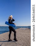 Woman Yoga Mat Beach Workout Fitness - A woman in black athletic wear stands on a pier overlooking the sea, holding a blue yoga mat.
