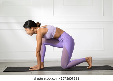 A woman in yoga pose on black mat, wearing purple activewear, practicing mindfulness and flexibility in bright indoor space with white walls