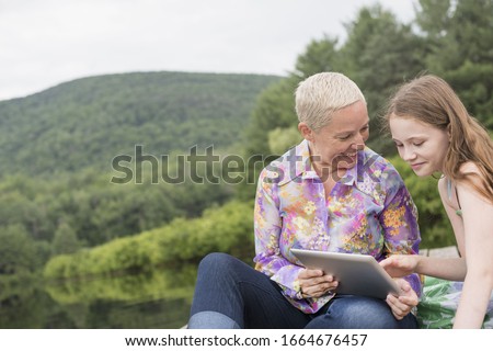 A woman and a young girl together on a lake dock using a digital tablet.