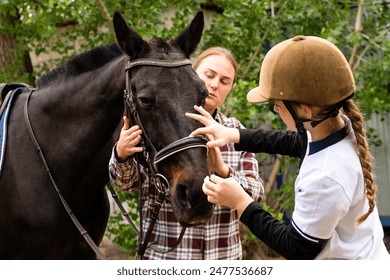 Woman and young rider girl carefully adjusting the bridle on a black horse. Equine-assisted learning. Therapeutic horseback riding. Hippotherapy - Powered by Shutterstock