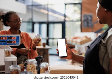 Woman in zero waste shop using mockup mobile device to analyze products. Vegan customer checking food item suitable for his diet while female vendor holds smartphone with isoalted white screen. - Powered by Shutterstock