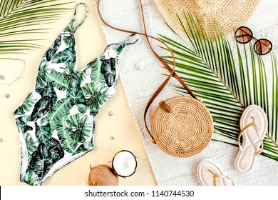 Woman's beach accessories: swimsuit with tropical print, rattan bag, straw hat, tropical palm leaves on yellow background. Summer background. Flat lay, top view.
