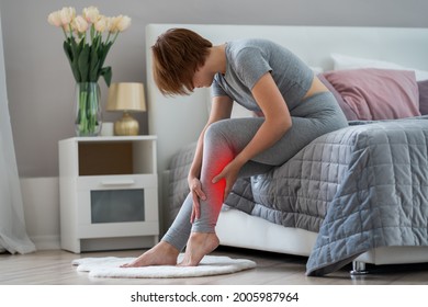 The woman's calf muscle cramped, massage of female leg in home interior, painful area highlighted in red - Shutterstock ID 2005987964