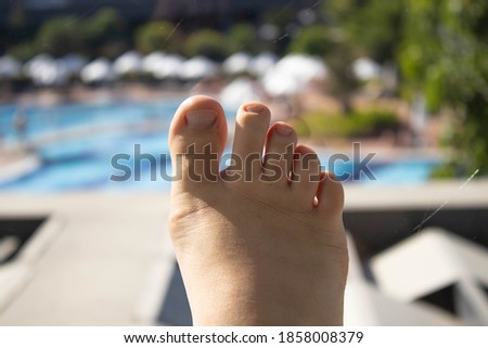 A woman's feet is in focus, at the balcony of a hotel room and a pool is behind. Vacation during the pandemic.