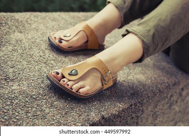 Woman's Feet In Yellow Stylish Summer Sandals With Dark Nail Polish, In Green Summer Pants Sitting On The Wall