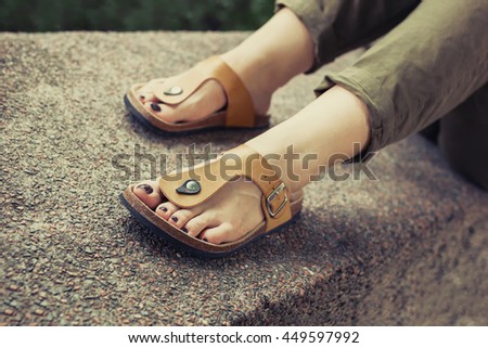 Woman's feet in yellow stylish summer sandals with dark nail polish, in green summer pants sitting on the wall