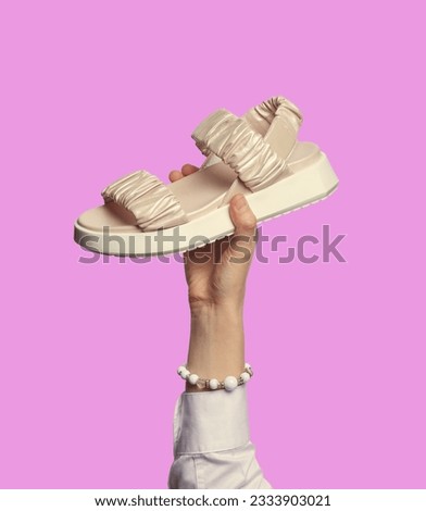 A woman's hand with a bracelet, wearing a white shirt, holding a stylish shiny beige sandal isolated on a bright lilac background. Graceful hand showcasing footwear