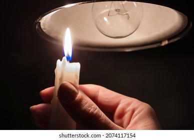 A Woman's Hand With Burning Candle In Complete Darkness Looking On A Switched Off Light Bulb At Home. Blackout, Electricity Off, Energy Crisis Or Power Outage, Concept Image.