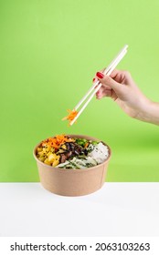 A woman's hand with chopsticks over a salad poke bowl and tuna, carrot, cucumber and pineapple with a green background. white floor - Shutterstock ID 2063103263