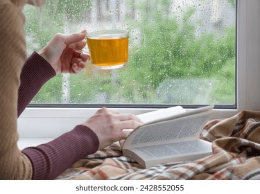 A woman's hand with a cup of tea. An open book and a soft blanket. The window glass is covered with raindrops. Springtime - Powered by Shutterstock