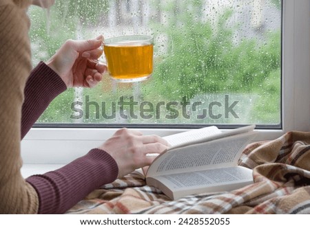 A woman's hand with a cup of tea. An open book and a soft blanket. The window glass is covered with raindrops. Springtime