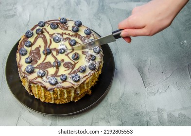 Woman's Hand Cuts A Large Homemade Cake With A Knife On The Background Of A Concrete Tabletop