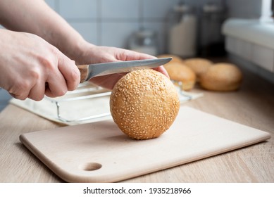 Woman's Hand Cutting A Sesame Seed Bun For A Hamburger With A Knife In The Kitchen, Cooking At Home, Close Up.