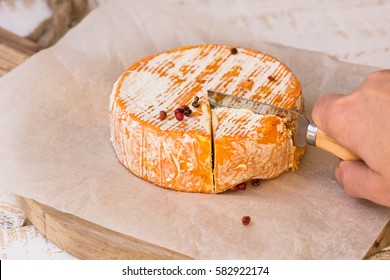 Woman's hand cutting whole wheel of soft French, German cheese with orange rind with mold on parchment paper, wood cutting board, rustic kitchen interior - Powered by Shutterstock