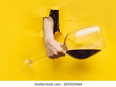 A Woman's Hand Emerges Through A Torn Hole In Yellow Paper With A Large Glass Of Red Wine. The Concept Of Alcoholism, Drunkenness And Hangover.