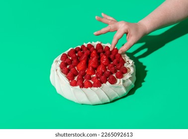 Woman's hand grabbing a raspberry from a pavlova cake, minimalist on a green background. Homemade pavlova cake with whipped cream filling, strawberries, and raspberries on top of it. - Powered by Shutterstock