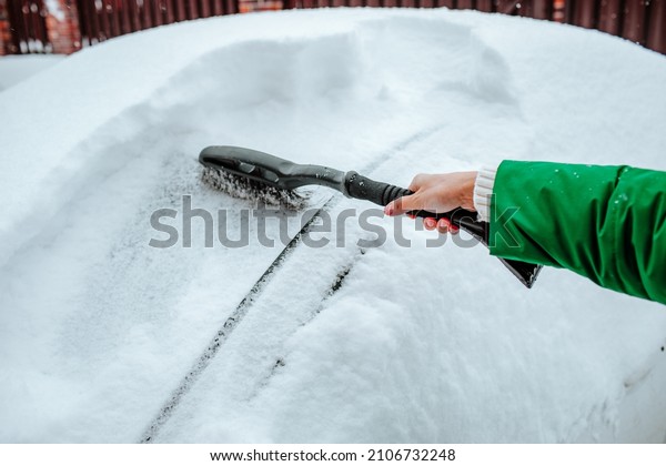 Woman\'s hand in green winter\
coat cleaning snowing car with the brush in the morning. Winter\
time.