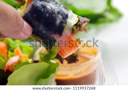Woman's hand hold roll of seaweed wrapping salad with intimate crab stick dipping in spicy sauce with miso or bean paste with chili in small bowl, blurred green vegetable, white isolated background 