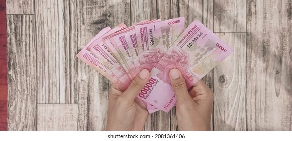 Woman's hand holding 10 pieces of Indonesian banknotes worth 100,000 rupiah, on a wooden paper background. Holding by spreading Indonesian rupiah banknotes with women's hands - Shutterstock ID 2081361406