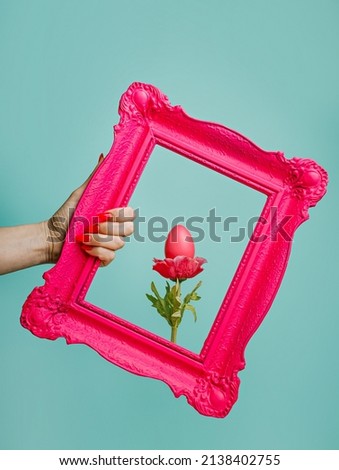 Woman's hand holding antique pink frame with marigold flower and colored egg against vibrant turquoise background. Creative Easter holiday concept. Abstract and surreal composition.