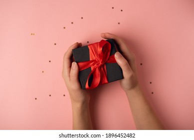 Woman's Hand Holding Black Gift Box With Red Ribbon On The Rose Background. The Concept Of Valentine's Day.