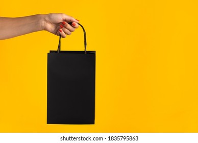 Woman's Hand Holding Black Paper Shopping Bag In Front Of Yellow Background. Black Friday Concept