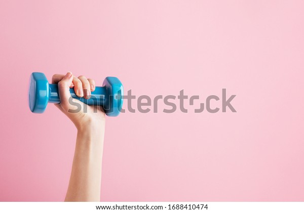 Womans hand holding\
blue dumbbell isolated on pink background. Equipment for home\
workout. Fitness and activity. Sport and healthy lifestyle concept.\
Copy space in right side