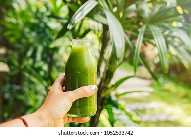 Woman's hand holding a bottle with green cold-pressed juice, nature background. Healthy eating, detoxing, juicing, fasting, body cleancing concept