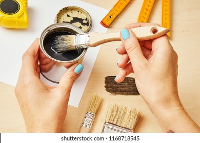 woman's hand holding a brush and dips in a can of paint on a wooden table