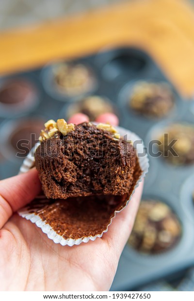 Woman\'s hand holding eaten or bitten healthy gluten free\
chocolate muffin with walnuts. Homemade, freshly baked goods with\
cocoa powder and dark chocolate placed in a muffin tray in\
background. 