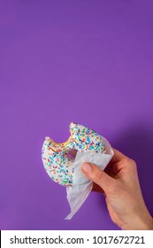 Woman's Hand Is Holding A Half Eaten Bagel In Front Of Purple Background. Sweetness And Happiness Concept. 