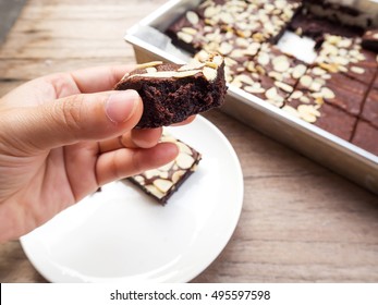 Woman's Hand Holding Homemade Sliced Brownie. Eaten Brownie. With Brownies  In Baking Tray And White Plate Are Background. Favorite Dessert For Chocolate Lover. Brownies With Almond Sliced Topping.