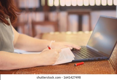woman's hand holding pen marking on proofreading paper with laptop on desk in office 
