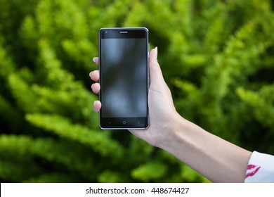 Woman's Hand Holding A Phone With A Large Screen Against  Background Of Green Bushes