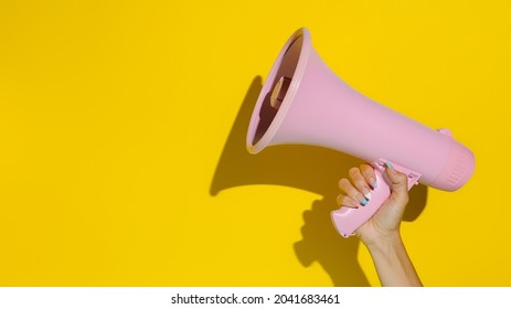 A woman's hand holding a pink megaphone isolated on a yellow background. Creative announcement concept. Loud voice of women. Women's rights and voice. Advertisement mock up with copy space for text.