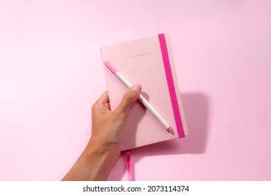 woman's hand holding a pink notebook with pen on pink background