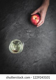 Woman's Hand Holding Red Apple And Glass Of White Wine On Dark Background. Overhead Shot.