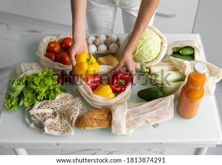 Woman's hand, holding a reusable grocery bag with vegetables on a kitchen at home and takes pepper out. Zero waste and plastic free concept. Mesh cotton shopper with vegetables. Ecology.