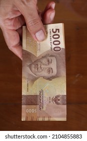 Woman's hand holding and showing stack of five thousand rupiahs paper moneys isolated on dark brown wood textured background. Vertically captured giving several of RP 5000 new paper Indonesia money. 