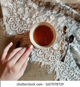 Woman's Hand Holding Small Vintage Teacup On Lace And Wood Background, Flat Lay Composition With A Tea Cup And Two Tea Spoons, Dark Black Chinese Tea Essence In A Small Traditional Ceramic Tea Cup.