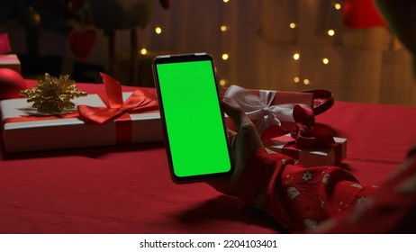 Woman's Hand Holding Smartphone With Green Screen Chroma Key. Woman On The Background Of A Home Interior Decorated On New Years Eve. Online Communication Via Video Chat. Christmas Holidays Concept.