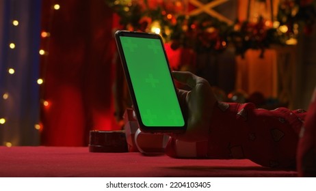 Woman's Hand Holding Smartphone With Green Screen Chroma Key. Woman On The Background Of A Home Interior Decorated On New Years Eve. Online Communication Via Video Chat. Christmas Holidays Concept.